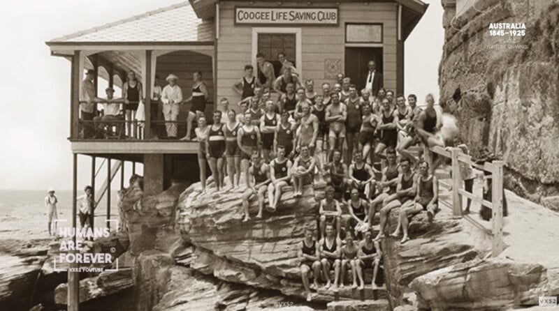 A large group of people in vintage swimsuits pose on rocks outside the Coogee Life Saving Club building, perched above the ocean in Australia, circa 1925.