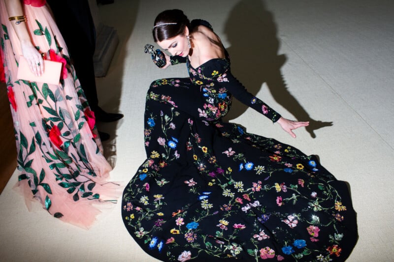 A woman in a long, black, off-the-shoulder gown with colorful floral designs sits gracefully on the floor, her hand supporting her. Another person in a light pink floral dress stands nearby, holding a pale clutch.