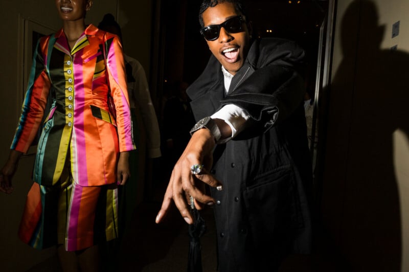 A man in a black coat and sunglasses smiles and gestures toward the camera, while a woman in a brightly colored striped suit stands nearby. The scene is lively and well-lit.