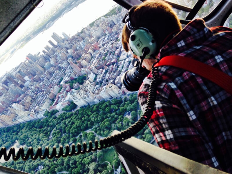 A photographer wearing headphones leans out of a helicopter, taking pictures of a city skyline and a large park below. The photo is taken at an angle, showing tall buildings and green tree-filled spaces.