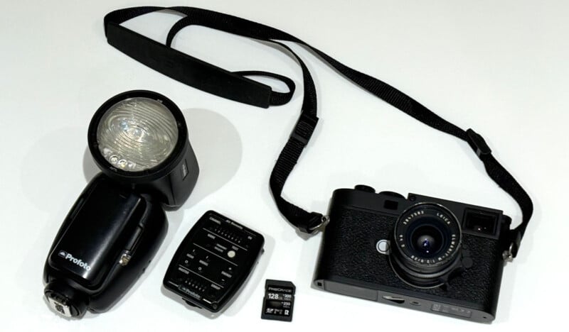 A digital camera with a lens and strap, an external flash, a wireless flash trigger, and a 128GB SD memory card are arranged on a white surface.
