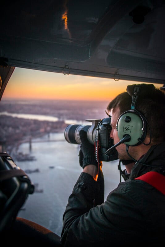 A person wearing headphones and gloves holds a camera, taking a photo from inside a helicopter flying over a city and river at sunset.
