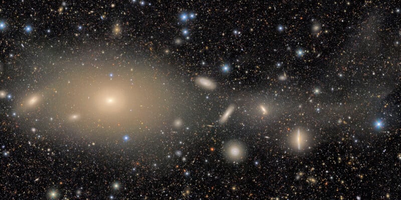 A dense star field in deep space shows glowing galaxies, clusters of stars, and cosmic dust set against a dark background, with several large, bright elliptical galaxies standing out prominently.