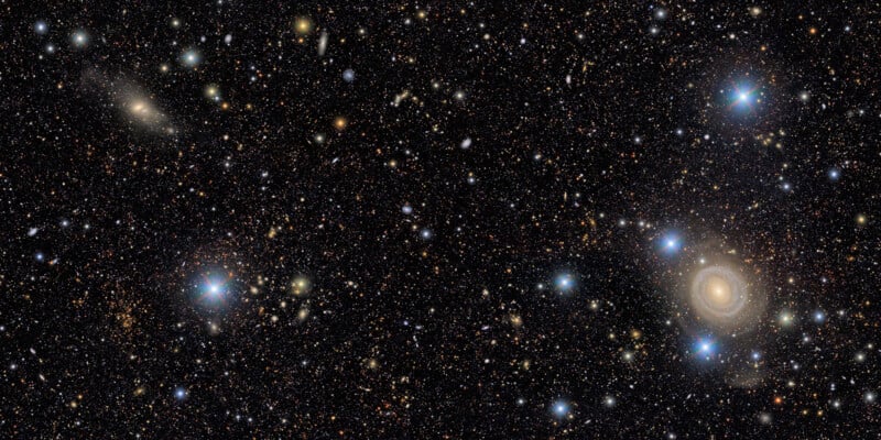 A dense star field in deep space, showing countless stars, distant galaxies, and a large spiral galaxy glowing brightly on the right amid sparkling blue and white stars against a dark backdrop.