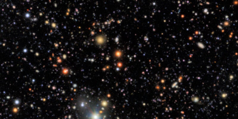 A view of deep space showing countless stars and galaxies scattered across a dark sky, with bright spots in various colors including white, orange, and yellow.