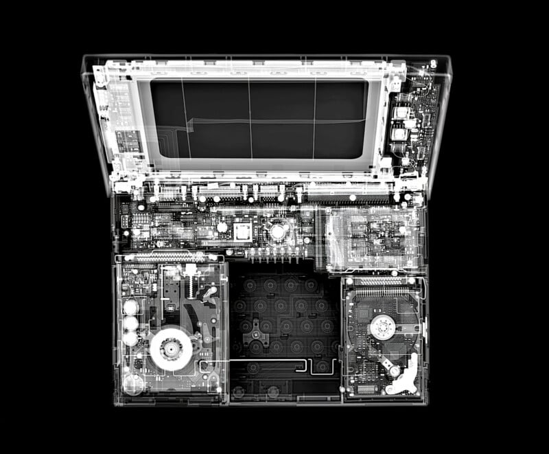 X-ray image of an open laptop viewed from above, revealing internal components like the motherboard, hard drive, cooling fan, and circuits, with the outline of the screen and keyboard visible.