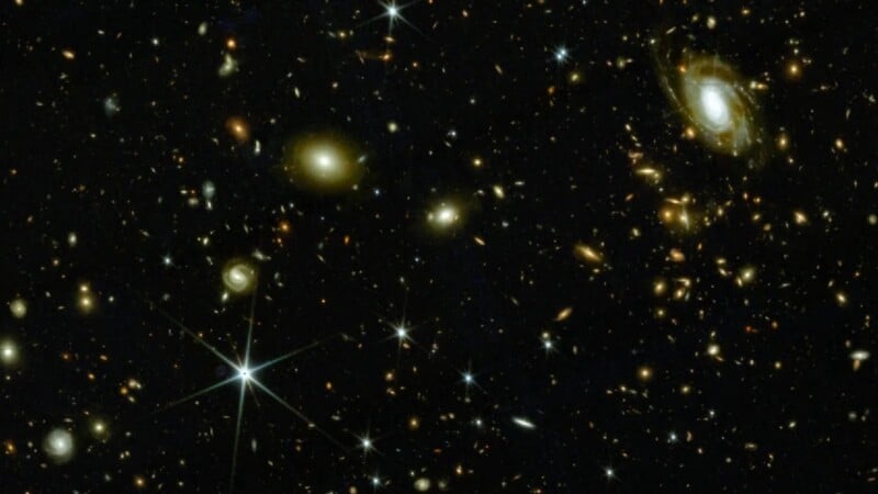 A deep space image shows numerous galaxies of various shapes and sizes scattered across a dark background, with some bright stars and spiral galaxies visible among distant cosmic objects.