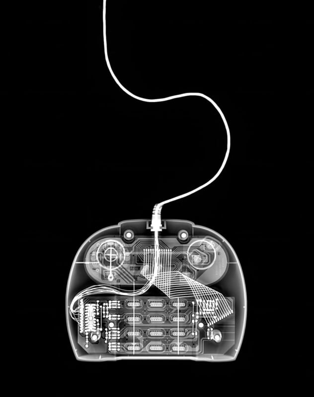X-ray image of a video game controller with a cable, showing internal electronic components, wiring, circuit board, and buttons against a black background.