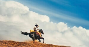 A person wearing traditional clothing rides a galloping horse across a dusty, grassy hill with dramatic white clouds and a blue sky in the background.