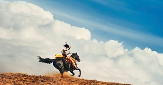 A person wearing traditional clothing rides a galloping horse across a dusty, grassy hill with dramatic white clouds and a blue sky in the background.
