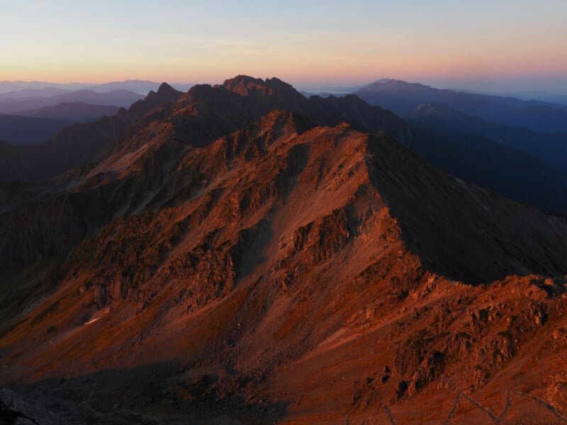 Rugged mountain ridge bathed in warm orange sunlight during sunrise or sunset, with distant peaks and valleys visible under a clear sky.