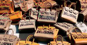 A pile of luxury handbags in various shades of brown and tan, each with the word "DELUSIONAL" spray-painted in black across the front.