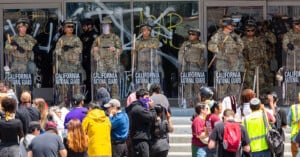 A line of California National Guard troops in riot gear stands in front of a building with glass doors covered in graffiti, facing a crowd of protesters gathered on the steps.
