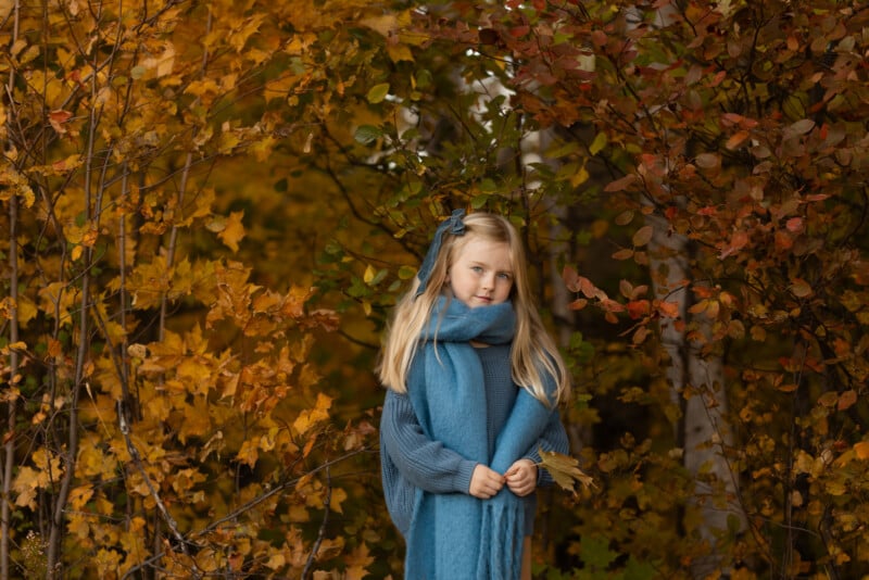 A young girl with long blonde hair, wearing a blue scarf and sweater, stands among autumn trees with yellow and orange leaves. She holds a few leaves and looks at the camera.