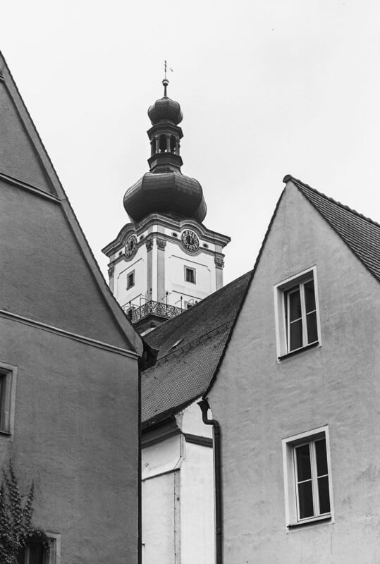 A black and white photo of a church tower with a dome-shaped roof rising above nearby buildings with sloped roofs and windows. The sky is overcast.