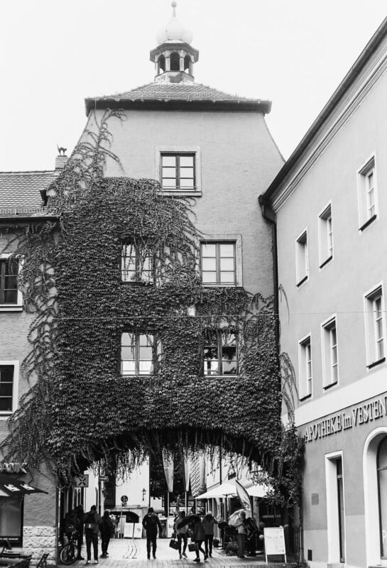 Black and white photo of a tall building with ivy climbing its facade, arched passageway at the base, and people walking underneath. Signs on nearby buildings are visible, one reads "APOTHEKE IM VESTEN.