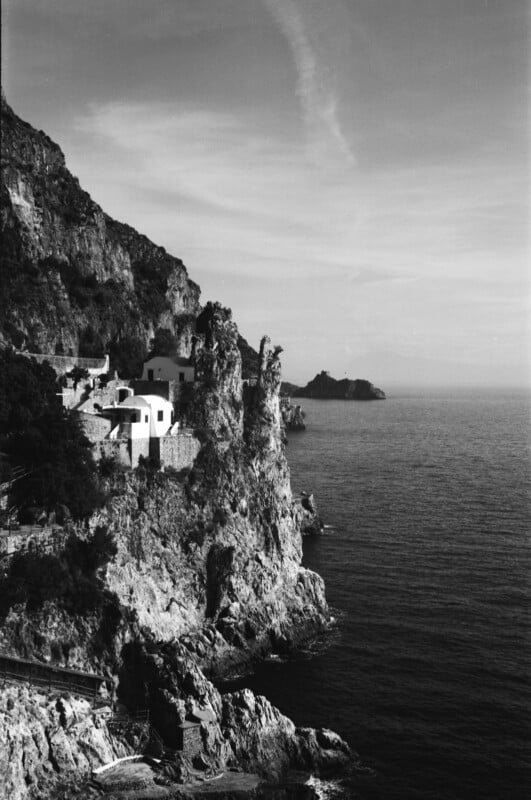 Black and white photo of houses perched on rocky cliffs overlooking a calm sea, with jagged rocks jutting out and steep hills rising in the background under a lightly clouded sky.