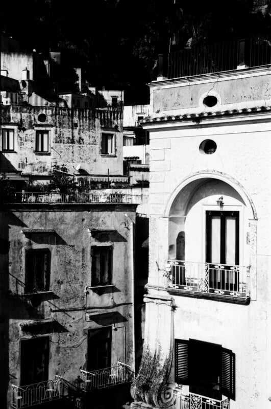 Black and white photo of old apartment buildings with balconies and arched windows. The sunlight casts sharp shadows, highlighting the textured walls and architectural details.
