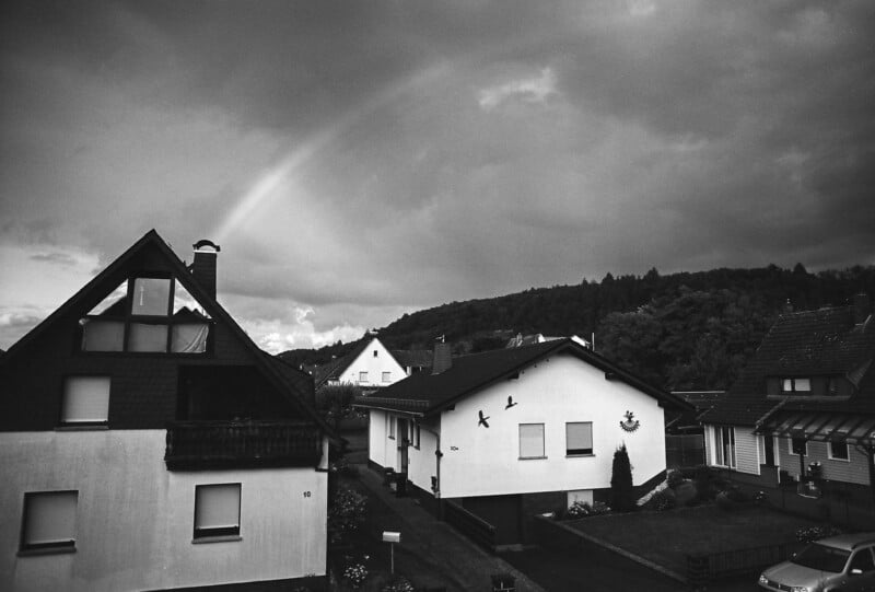 A black and white photo shows suburban houses under a cloudy sky, with a faint rainbow visible. Trees line the background, and cars are parked near the houses, creating a calm, quiet neighborhood scene.