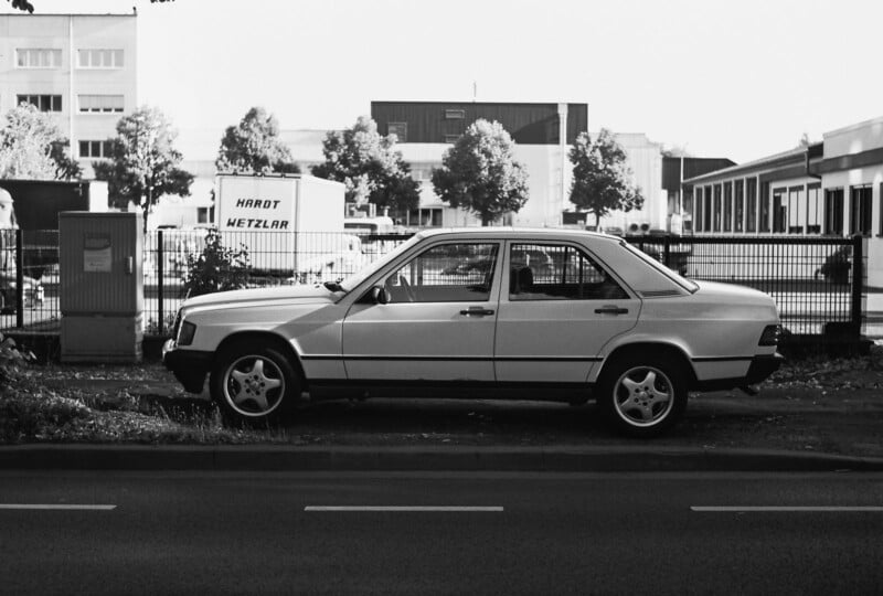A white sedan is parked on the side of a street next to a fence, with industrial buildings and trees in the background. The image is in black and white.