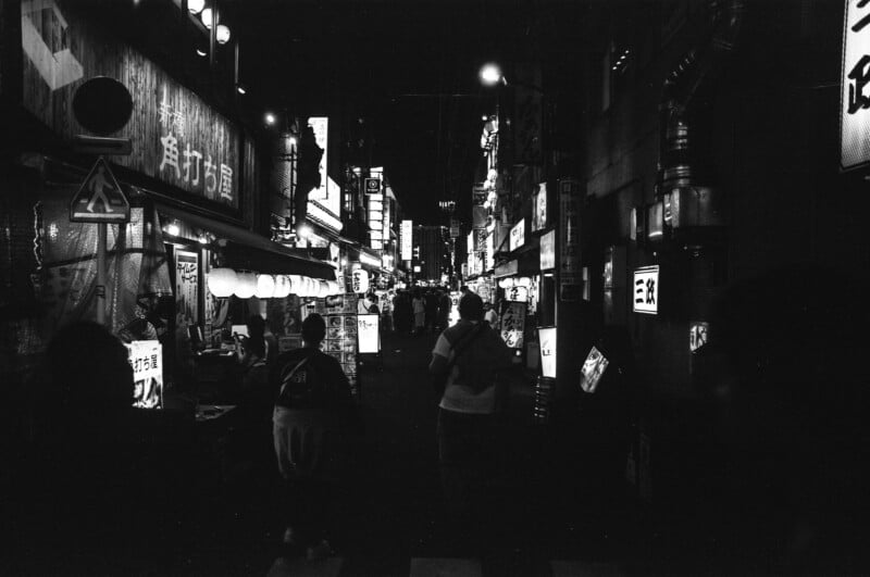 A black-and-white photo of a bustling city street at night, featuring glowing neon signs in Japanese, paper lanterns, and silhouettes of people walking along crowded sidewalks.