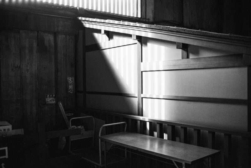 A beam of sunlight streams through a window, casting shadows and patterns on the wooden walls, a bench, and a table in a quiet, empty room with traditional Japanese architecture.