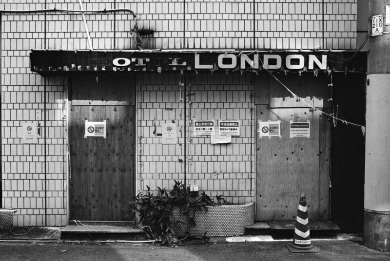 Black-and-white photo of a boarded-up building with a worn sign reading "HOTEL LONDON." Notices are posted on the doors, plants and a traffic cone sit in front, and the exterior is tiled.