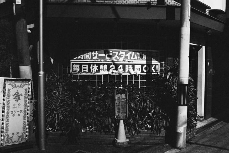 A black and white photo of a Japanese street scene, featuring a sign with Japanese text behind a fence, surrounded by plants and partially obscured by shadows, with poles and a patterned board in the foreground.