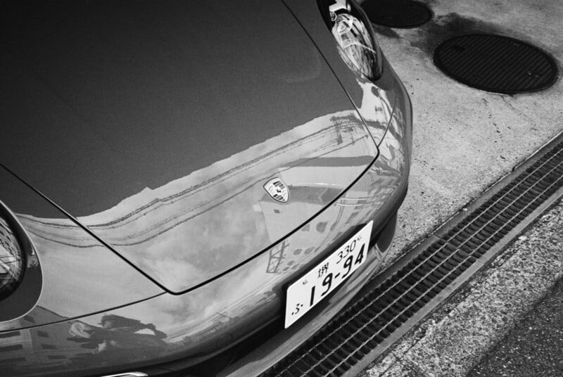 A black-and-white photo shows the hood of a Porsche with reflections of buildings and clouds. A Japanese license plate and part of the street are visible beside the car.