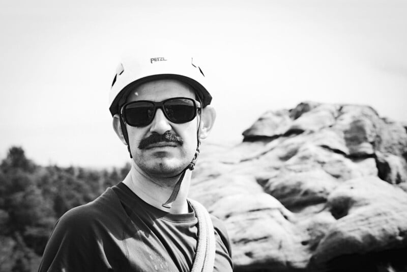 A man wearing sunglasses, a helmet, and a climbing harness stands outdoors near large rocks. The photo is in black and white. Trees are visible in the background.