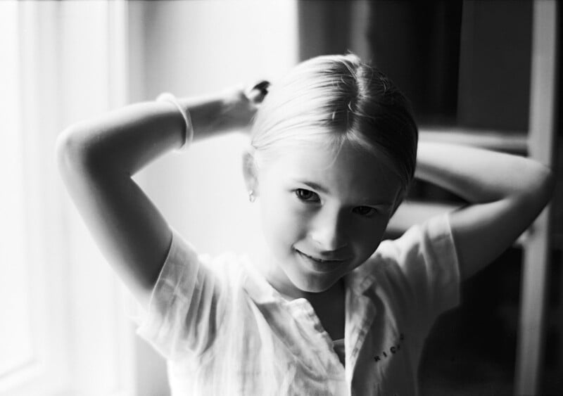 Black and white photo of a young girl with light hair, smiling as she pulls her hair back with both hands. She is wearing a light shirt and a bracelet, standing near a bright window.