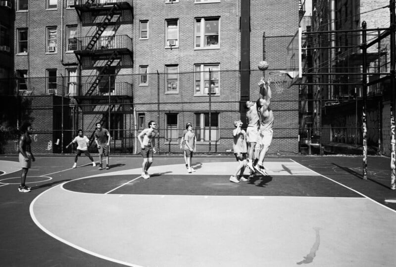 A group of men play basketball on an outdoor urban court, surrounded by tall brick apartment buildings with fire escapes. Two players jump beneath the hoop, reaching for the ball, while others watch or run nearby.