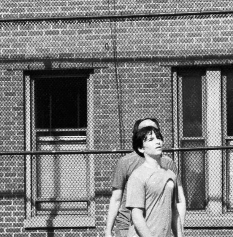 Two people stand close together outside in front of a brick building with barred windows, looking upward. The scene is in black and white, and a wire fence is visible behind them.