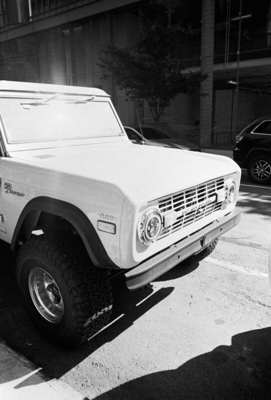 Black and white photo of a classic Ford Bronco parked on a city street, partially in sunlight, with a tree, glass building, and another vehicle in the background.