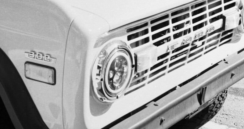 Close-up black and white photo of the front of a classic Ford truck, showing the round headlight, front grille with “FORD” lettering, and a “302” badge on the side.