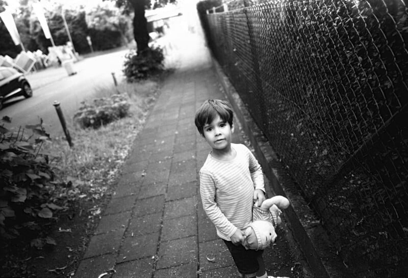 A young child stands on a sidewalk, holding a stuffed animal. The scene is in black and white, with a fence on one side and plants on the other. The child looks toward the camera with a neutral expression.
