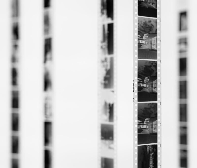 Black-and-white photo showing several strips of photographic film negatives hanging vertically. The image focuses on one strip while others are blurred in the background, creating a sense of depth.