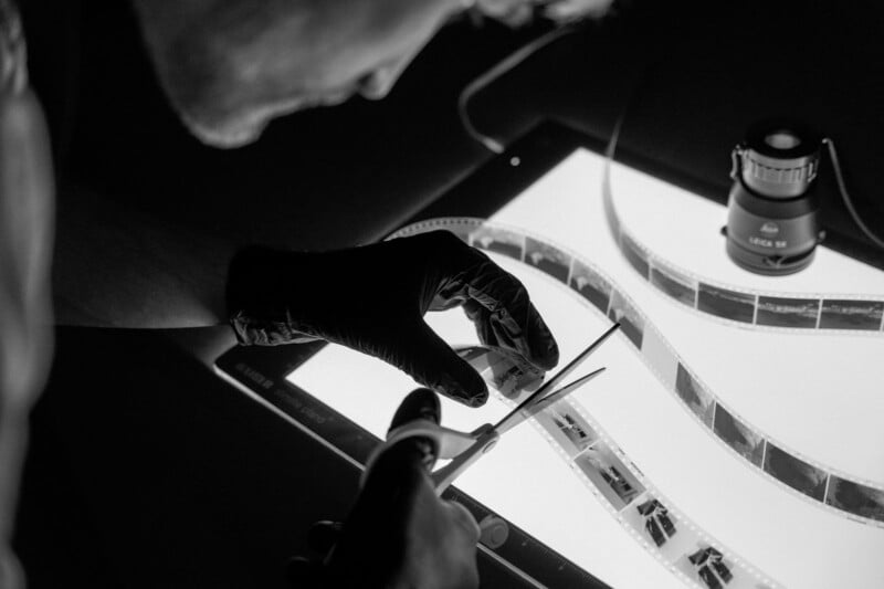 A person wearing black gloves uses scissors to cut a strip of photographic film negatives on a light table, with a magnifying tool nearby. The scene is in black and white.