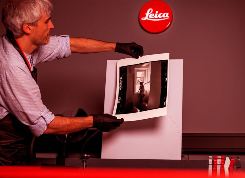 A man wearing gloves holds up a black and white photo print in a darkroom. A Leica logo is illuminated in red on the wall above him, with photographic tools and trays visible on the table.