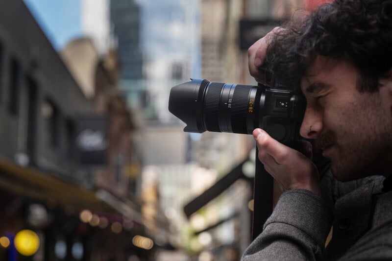 A man with curly hair and a mustache is holding a camera up to his eye, taking a photo on a city street with blurred buildings and lights in the background.