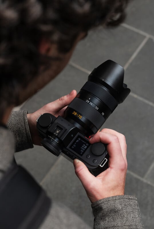 A person with curly hair holds a digital camera with a large zoom lens, adjusting camera settings on the screen while standing on gray tile flooring.