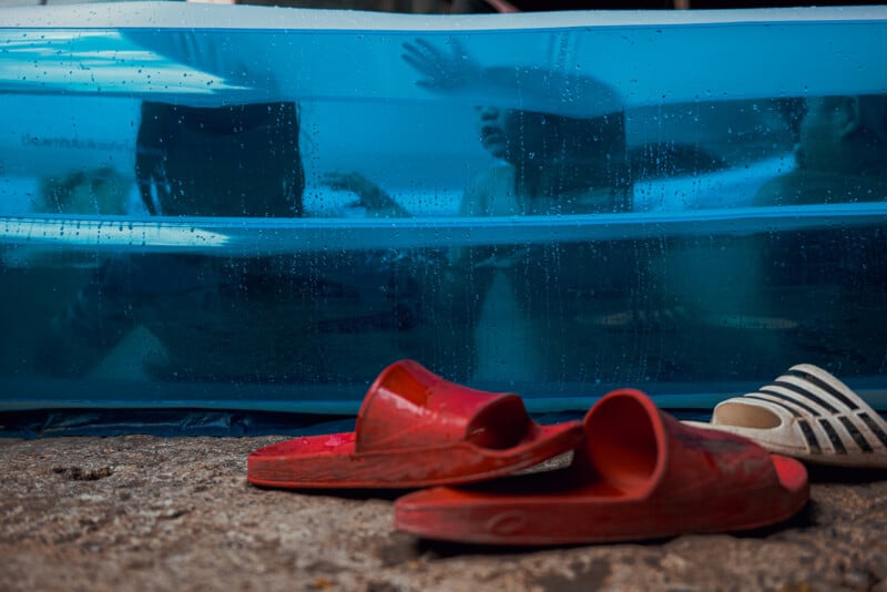 Two children are seen through the blue side of an inflatable pool, pressing their hands against it. In front of the pool on the ground, there are a pair of red sandals and one white striped sandal.