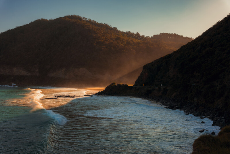 Waves crash against a rocky coastline at sunset, with sunlight streaming over tree-covered hills and illuminating mist rising from the shore.