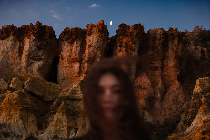 A blurred woman with hair covering her face stands in front of dramatic, sunlit rocky cliffs at dusk, with a crescent moon visible in the darkening sky above.