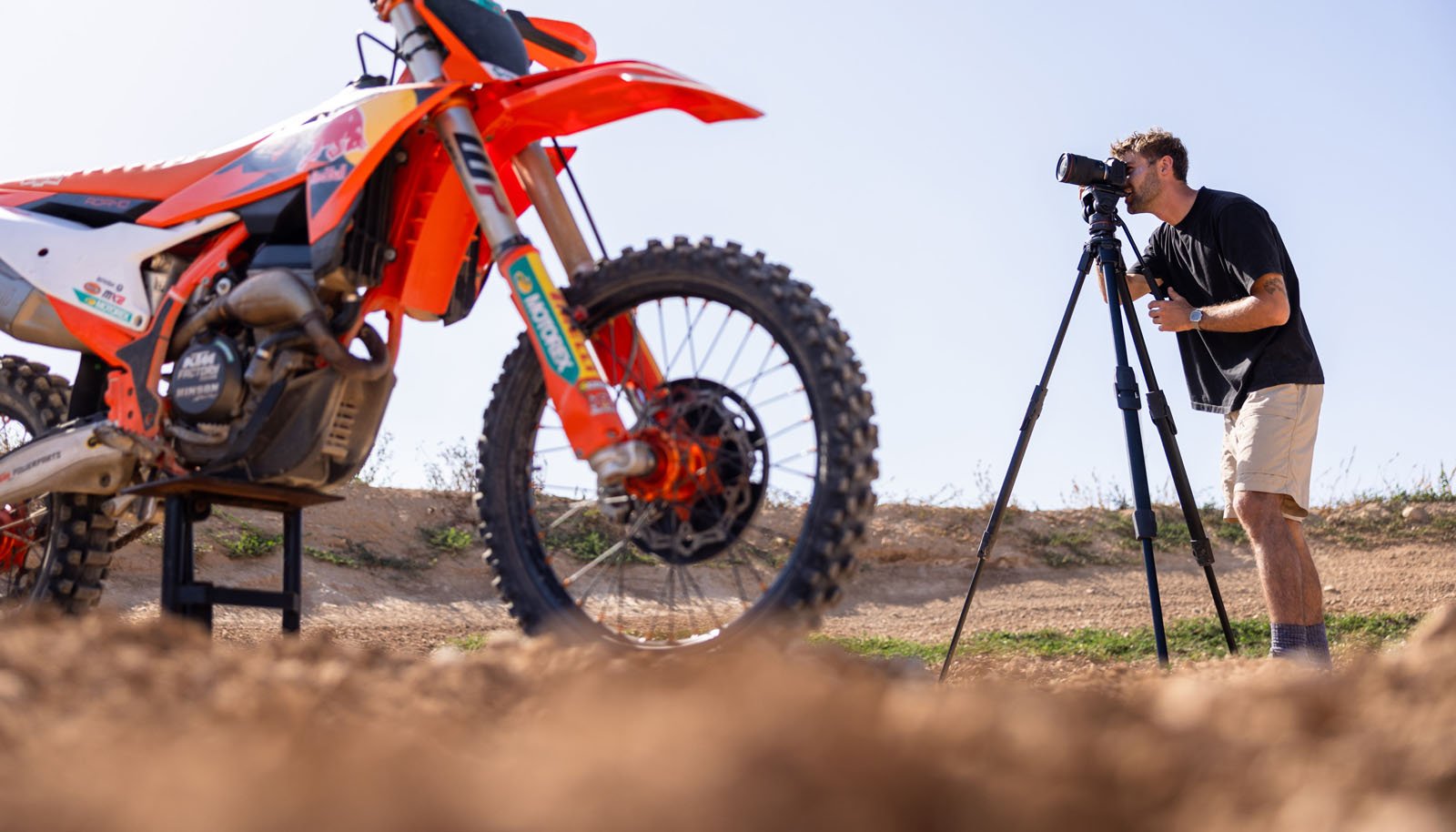 A man stands outdoors on dirt, taking photos with a camera on a tripod, focusing on a parked orange dirt bike in the foreground. The sky is clear and sunny.
