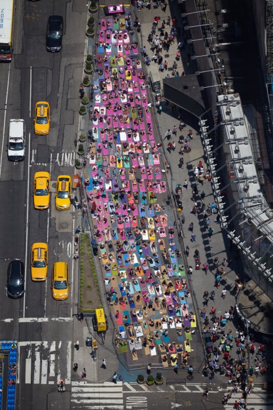 Aerial view of colorful yoga mats with people practicing yoga on a blocked-off street in a busy city, surrounded by yellow taxis and tall buildings.