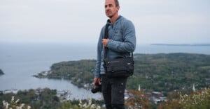 A man in a blue jacket stands on a hill overlooking a coastal landscape with trees, water, and buildings below, holding a camera and wearing a black shoulder bag.