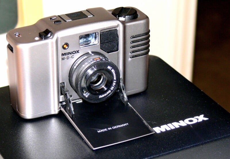 A Minox MDC compact camera with a lens and an open panel labeled "Made in Germany," sits on a black surface with the Minox logo visible.