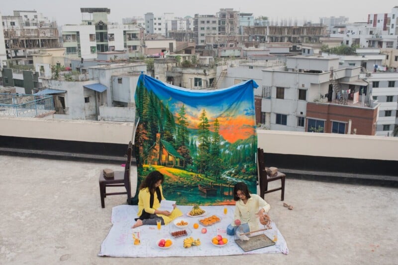 Two people have a picnic on a rooftop, sitting on a blanket with food and books. Behind them is a large tapestry with a colorful nature scene, and city buildings are visible in the background.