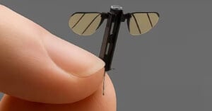 A close-up of a person’s fingers holding a tiny robotic insect with translucent wings and a thin body, resembling a small mechanical fly or drone, against a plain gray background.